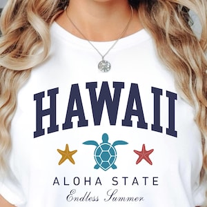 Peut inclure: T-shirt blanc avec le mot "HAWAII" en bleu marine en arc, une étoile de mer dorée, une tortue turquoise et une étoile de mer rouge. Les mots "ALOHA STATE" et "Endless Summer" sont en dessous.