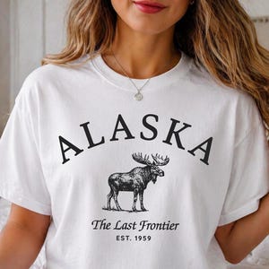 Pode incluir: Camiseta branca com a palavra "ALASKA" em arco e uma ilustração detalhada de um alce abaixo. O texto "The Last Frontier" e "EST. 1959" são impressos abaixo. A camiseta é de estilo casual.