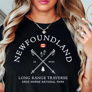 Peut inclure: T-shirt noir avec texte et graphiques blancs. Le texte "NEWFOUNDLAND" est au-dessus de bâtons de randonnée croisés et d'un drapeau canadien. Il est écrit "LONG RANGE TRAVERSE GROS MORNE NATIONAL PARK" et "23 MILES".