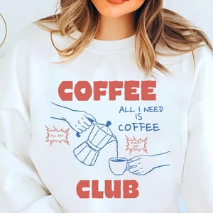 Peut inclure: Sweat-shirt blanc avec un motif rouge et bleu sur le thème du café. Le motif comprend les mots "COFFEE CLUB" et "ALL I NEED IS COFFEE", ainsi qu'une illustration de café versé. Le sweat-shirt porte également le texte "Atlas Doll Crafts" dans un cercle doré.