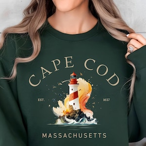 Peut inclure: Sweat-shirt vert forêt avec un motif de phare de Cape Cod. Le motif présente un phare rouge et blanc avec les mots "CAPE COD MASSACHUSETTS" et l'année "1637". Le sweat-shirt a un col rond.