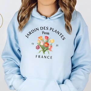 Peut inclure: Un sweat à capuche bleu clair avec un imprimé floral et le texte "JARDIN DES PLANTES Paris FRANCE". Le motif comprend un bouquet de tulipes colorées. Le sweat à capuche a une capuche et une poche avant. Le logo "Atlas Dali Crafts" est dans un cercle doré.