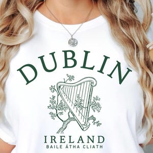 May include: White t-shirt with a green graphic of a harp and the word "DUBLIN" arched above it. Below the harp, the text reads "IRELAND" and "BAILE ÁTHA CLIATH". The shirt is a casual style.