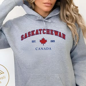 May include: A light grey hooded sweatshirt with a front pocket. The sweatshirt features the word "SASKATCHEWAN" in red arched lettering, with "EST. 1905" and "CANADA" below a red maple leaf. The words "Sport grey" are printed on the bottom right.