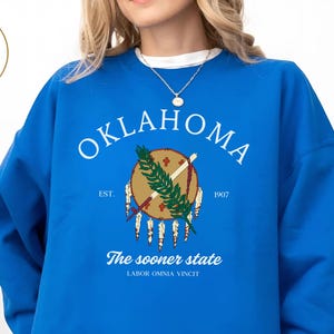 Peut inclure: Un sweat-shirt bleu roi avec le mot "OKLAHOMA" en blanc. En dessous, un emblème circulaire comprend un bouclier, des plumes et le texte "The sooner state". Le sweat-shirt a un col rond. Le texte "LABOR OMNIA VINCIT" est également visible.