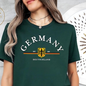 Puede incluir: Camiseta verde oscuro con la palabra "GERMANY" en letras blancas arqueadas. Debajo, un emblema amarillo, negro y rojo con el texto "EST. 1871" y "DEUTSCHLAND". La camiseta es de cuello redondo.