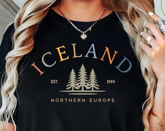 May include: Black t-shirt with the word "ICELAND" in colorful letters, above a graphic of three trees and the text "EST. 1944 NORTHERN EUROPE". The shirt is a crew neck.
