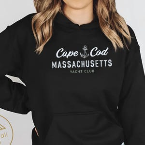 Op de afbeelding: Zwarte hoodie met de witte tekst "Cape Cod MASSACHUSETTS YACHT CLUB". Het ontwerp bevat een ankerafbeelding. De hoodie heeft een voorzak en een capuchon met trekkoord. Het Atlas Dali Crafts-logo staat linksonder.