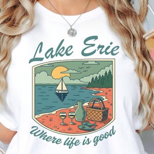 Peut inclure: T-shirt blanc avec une illustration de Lake Érié, représentant un voilier, le soleil et un pique-nique. Le texte "Lake Erie" et "Where life is good" sont affichés au-dessus et en dessous de l'illustration. Le t-shirt a également le texte "Atlas Dall Crafts" en bas.