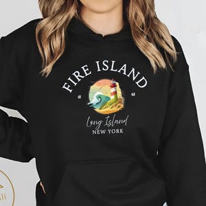 Può includere: Felpa con cappuccio nera con una grafica circolare con la scritta "Fire Island Long Island New York". Il disegno mostra un faro, onde e sabbia. La felpa ha una tasca frontale e un cappuccio con coulisse.