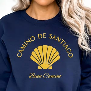 May include: Navy blue sweatshirt with the words "CAMINO DE SANTIAGO" and "Buen Camino" in gold lettering. A gold scallop shell design is centered below the text. The sweatshirt is a crew neck style.
