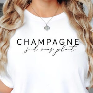 Peut inclure: T-shirt blanc avec l'inscription noire "CHAMPAGNE s'il vous plait". Un verre transparent avec un motif floral et une paille est tenu à la main. Le t-shirt a un col rond et est fait d'une matière douce.