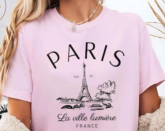 Op de afbeelding: Een lichtroze t-shirt met het woord "PARIS" in zwarte letters. Daaronder een zwarte illustratie van de Eiffeltoren en de tekst "La ville lumière FRANCE". Het shirt heeft korte mouwen.