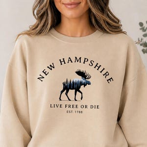 Peut inclure: Un sweat-shirt à col rond couleur sable avec un motif noir représentant un élan dont le corps contient une silhouette de forêt. Le texte NEW HAMPSHIRE, LIVE FREE OR DIE et EST. 1788 est imprimé en noir avec une police serif sur la poitrine.