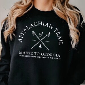 Peut inclure: Sweat-shirt noir avec l'inscription "APPALACHIAN TRAIL" en demi-cercle, avec des bâtons de randonnée croisés et le texte "MAINE TO GEORGIA". Le sweat-shirt indique également "THE LONGEST HIKING-ONLY TRAIL IN THE WORLD".
