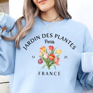 Peut inclure: Sweat-shirt bleu clair avec l'inscription "JARDIN DES PLANTES PARIS FRANCE" et un motif floral. Le sweat-shirt porte également les chiffres "19" et "26". Les mots "Light Blue" sont imprimés dans le coin supérieur droit.