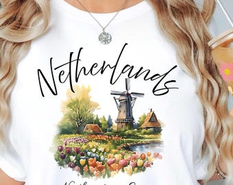 May include: White t-shirt with a watercolour illustration of a Dutch windmill, flowers, and the word "Netherlands." The text "Northwestern Europe Est. 1871" is below the image. The shirt also has the brand logo "Atlas Dali Crafts."
