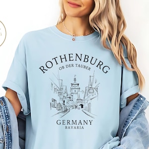 Può includere: T-shirt azzurra con grafica nera di Rothenburg ob der Tauber, Germania. Il design include il nome della città, un'illustrazione dettagliata degli edifici e la scritta "Germany Bavaria". La maglietta è in stile casual.
