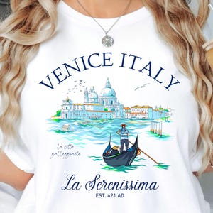 Peut inclure: T-shirt blanc avec un motif de Venise, Italie. Le graphisme comprend le texte "Venice Italy" au-dessus d'une illustration détaillée d'une scène vénitienne avec une gondole, des bâtiments et de l'eau. Le texte "La Serenissima" est en dessous.