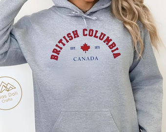 Puede incluir: Una sudadera con capucha gris claro con un bolsillo delantero y una capucha. La parte delantera presenta las palabras "British Columbia" en rojo, arqueadas sobre una hoja de arce roja y la palabra "Canadá" en azul. La sudadera es "Sport grey".