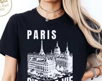 Puede incluir: Camiseta negra con un gráfico blanco de monumentos parisinos, incluyendo la Torre Eiffel y el Sacré-Cœur. Las palabras "PARIS" y "C'EST LA VIE" están impresas en blanco. La camiseta es de cuello redondo.