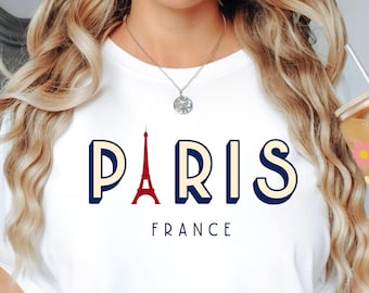 Op de afbeelding: Wit T-shirt met het woord "PARIS" in marineblauw en crème letters, met een rode Eiffeltoren-afbeelding. Het woord "FRANCE" staat onder het woord "PARIS". Een helder glas met een bloemenontwerp wordt in de hand gehouden.