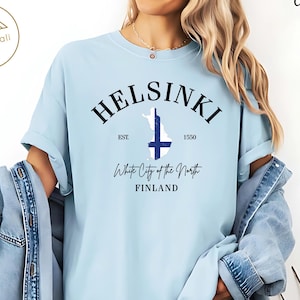 Peut inclure: T-shirt bleu clair avec le mot "Helsinki" en noir, une carte de la Finlande avec une croix bleue et le texte "White City of the North, Finland". Le t-shirt a des manches retroussées et est fait d'une matière douce.