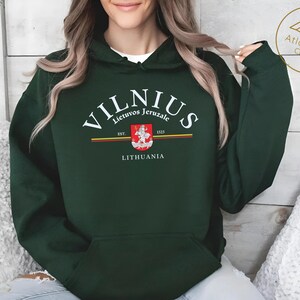 Könnte beinhalten: Dunkelgrüner Kapuzenpullover mit dem Wort "VILNIUS" in weißem, bogenförmigem Text. Darunter ein Wappen mit den Worten "Lietuvos Jeruzale" und "LITHUANIA". Der Pullover hat eine Vordertasche und eine Kapuze mit Kordelzug.