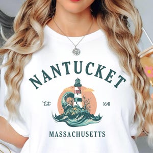 Peut inclure: T-shirt blanc avec un motif de phare et le texte "Nantucket Massachusetts" et l'année "1641". Le motif comprend un phare, des vagues et un coucher de soleil. Le t-shirt porte également le texte "Atlas Dall Crafts" en bas.