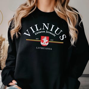 Könnte beinhalten: Schwarzes Sweatshirt mit dem weißen Wort "VILNIUS" in einem Bogen. Darunter sind die Texte "Lietuvos Jeruzale" und "LITHUANIA" zu sehen. Ein Wappen und der Text "EST. 1323" sind ebenfalls sichtbar. Das Sweatshirt hat einen Rundhalsausschnitt.