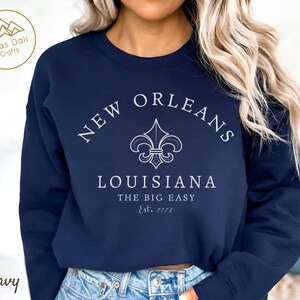 May include: Navy blue sweatshirt with white text and a fleur-de-lis design. The text reads "New Orleans Louisiana The Big Easy Est. 1718". The sweatshirt is a crew neck style.