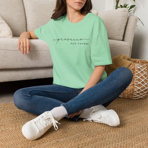 Op de afbeelding: Een lichtgroen T-shirt met korte mouwen en de zwarte tekst "prosecco PER FAVORE" op de borst gedrukt. Het shirt wordt gecombineerd met een donkerblauwe spijkerbroek en witte sneakers, wat zorgt voor een casual uitstraling met een eenvoudig ontwerp.