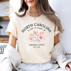 Może przedstawiać: Kremowa koszulka z napisem "NORTH CAROLINA", wsp&oacute;łrzędnymi i "TARHEEL STATE EST. 1789". Projekt zawiera r&oacute;żowe ilustracje kwiatowe. Koszulka jest w naturalnym kolorze.