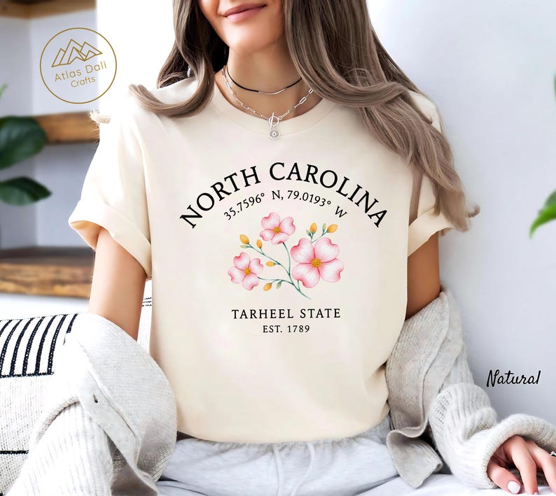 Może przedstawiać: Kremowa koszulka z napisem "NORTH CAROLINA", wsp&oacute;łrzędnymi i "TARHEEL STATE EST. 1789". Projekt zawiera r&oacute;żowe ilustracje kwiatowe. Koszulka jest w naturalnym kolorze.