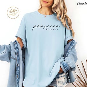 Peut inclure: T-shirt bleu clair avec l'inscription "prosecco PLEASE" en &eacute;criture noire. Le t-shirt est de style d&eacute;contract&eacute;. L'image montre &eacute;galement une veste en jean et le logo Atlas Dali Crafts.