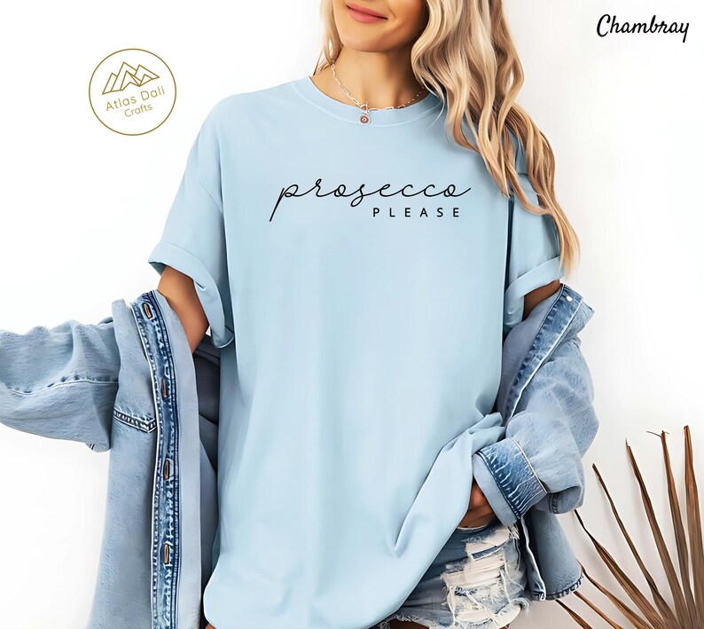 Peut inclure: T-shirt bleu clair avec l'inscription "prosecco PLEASE" en &eacute;criture noire. Le t-shirt est de style d&eacute;contract&eacute;. L'image montre &eacute;galement une veste en jean et le logo Atlas Dali Crafts.