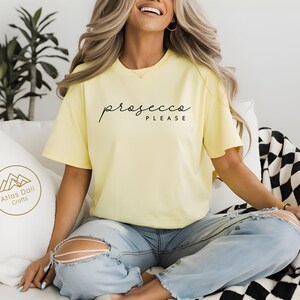 Peut inclure: T-shirt jaune clair avec l'inscription "prosecco PLEASE" en &eacute;criture noire. Le mod&egrave;le porte un jean bleu clair. Un oreiller blanc avec un logo dor&eacute; est &agrave; gauche. Une couverture &agrave; carreaux noirs et blancs est &agrave; droite.