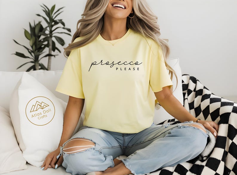 Peut inclure: T-shirt jaune clair avec l'inscription "prosecco PLEASE" en &eacute;criture noire. Le mod&egrave;le porte un jean bleu clair. Un oreiller blanc avec un logo dor&eacute; est &agrave; gauche. Une couverture &agrave; carreaux noirs et blancs est &agrave; droite.