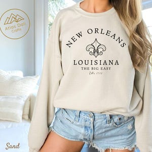 May include: Sand-colored sweatshirt with the text "NEW ORLEANS LOUISIANA THE BIG EASY Est. 1718" in black. The design includes a fleur-de-lis symbol. The sweatshirt is a crew neck style.