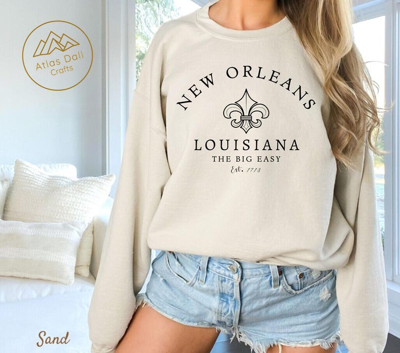 May include: Sand-colored sweatshirt with the text "NEW ORLEANS LOUISIANA THE BIG EASY Est. 1718" in black. The design includes a fleur-de-lis symbol. The sweatshirt is a crew neck style.