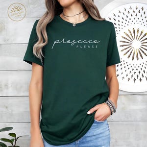 Peut inclure: Un t-shirt vert fonc&eacute; avec l'inscription "prosecco PLEASE" en &eacute;criture blanche. Le t-shirt est de couleur unie et a des manches courtes. Le mod&egrave;le porte un jean bleu et un collier.