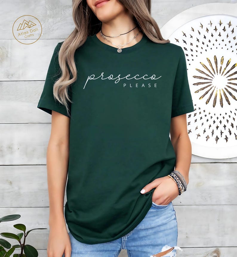 Peut inclure: Un t-shirt vert fonc&eacute; avec l'inscription "prosecco PLEASE" en &eacute;criture blanche. Le t-shirt est de couleur unie et a des manches courtes. Le mod&egrave;le porte un jean bleu et un collier.