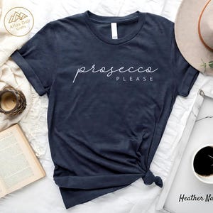Peut inclure: T-shirt bleu marine avec l'inscription "prosecco PLEASE" en &eacute;criture blanche. Le t-shirt est nou&eacute; en bas. Un chapeau, une tasse de caf&eacute;, une bougie et un livre sont en arri&egrave;re-plan.