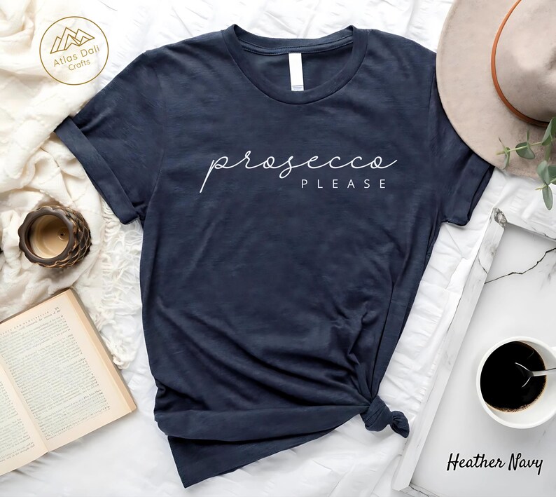 Peut inclure: T-shirt bleu marine avec l'inscription "prosecco PLEASE" en &eacute;criture blanche. Le t-shirt est nou&eacute; en bas. Un chapeau, une tasse de caf&eacute;, une bougie et un livre sont en arri&egrave;re-plan.