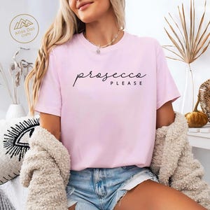 Peut inclure: Un t-shirt rose clair avec l'inscription "prosecco please" en &eacute;criture noire. Le t-shirt est &agrave; manches courtes et a un col rond. Le mod&egrave;le porte un short en jean bleu et un cardigan beige.