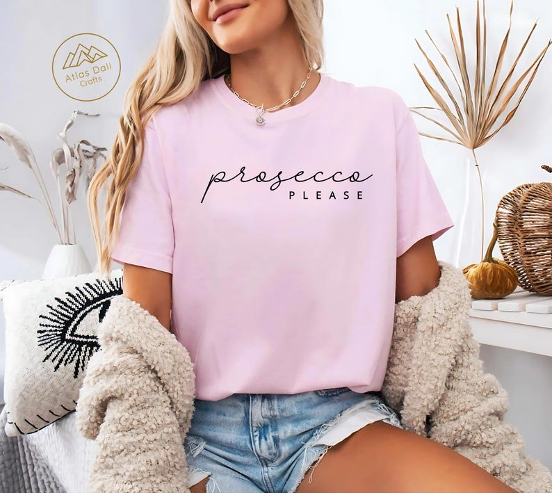 Peut inclure: Un t-shirt rose clair avec l'inscription "prosecco please" en &eacute;criture noire. Le t-shirt est &agrave; manches courtes et a un col rond. Le mod&egrave;le porte un short en jean bleu et un cardigan beige.