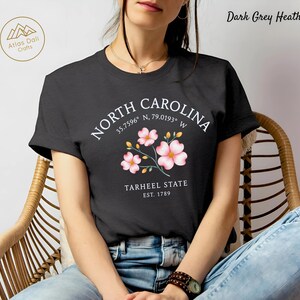 Może przedstawiać: Ciemnoszara koszulka z melanżem z napisem "NORTH CAROLINA", wsp&oacute;łrzędnymi i "TARHEEL STATE EST. 1789". Projekt zawiera r&oacute;żowe ilustracje kwiatowe. Koszulka ma okrągły dekolt.