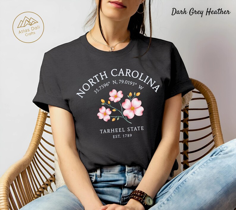 Może przedstawiać: Ciemnoszara koszulka z melanżem z napisem "NORTH CAROLINA", wsp&oacute;łrzędnymi i "TARHEEL STATE EST. 1789". Projekt zawiera r&oacute;żowe ilustracje kwiatowe. Koszulka ma okrągły dekolt.