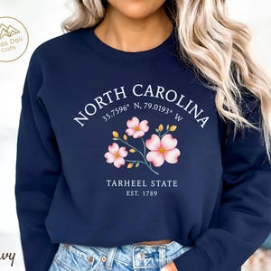 Op de afbeelding: Marineblauw sweatshirt met de tekst "NORTH CAROLINA", co&ouml;rdinaten en "TARHEEL STATE EST. 1789". Het heeft roze en gele bloemmotieven. De sweatshirt heeft een ronde hals en lange mouwen.