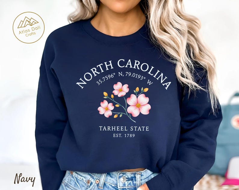 Op de afbeelding: Marineblauw sweatshirt met de tekst "NORTH CAROLINA", co&ouml;rdinaten en "TARHEEL STATE EST. 1789". Het heeft roze en gele bloemmotieven. De sweatshirt heeft een ronde hals en lange mouwen.